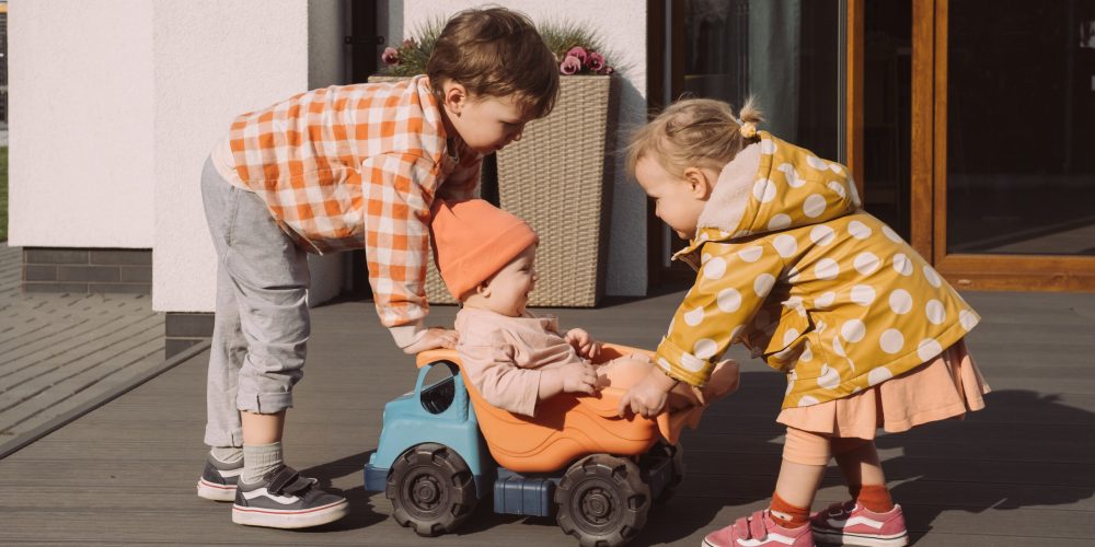 Brother sister giving a ride to baby sister in big toy car outdoors. Children having fun near house. Boy and girl racing with pleasure. Kids games of different age. Suburban life